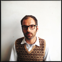 A man wearing glasses and a sweater vest sits in front of a white wall